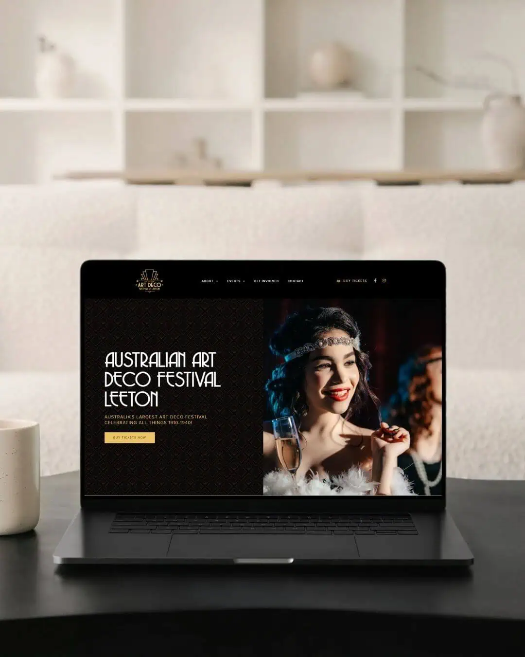 A laptop displaying a website for the Australian Art Deco Festival Leeton sits on a black table with a white mug in a modern living room.