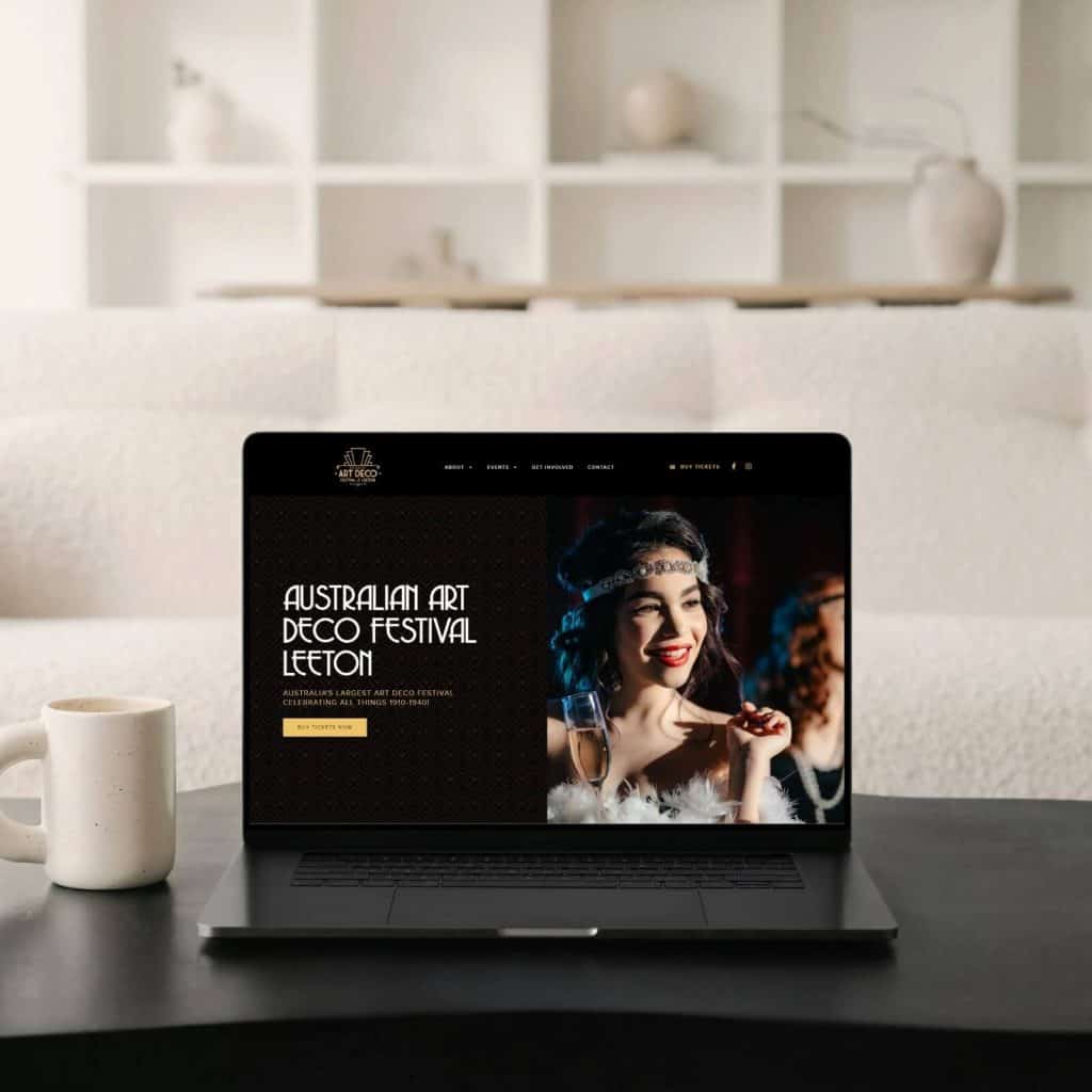 A laptop on a table displays an "Australian Art Deco Festival Leeton" webpage; a coffee mug sits beside it, with a white sofa and shelves in the background.