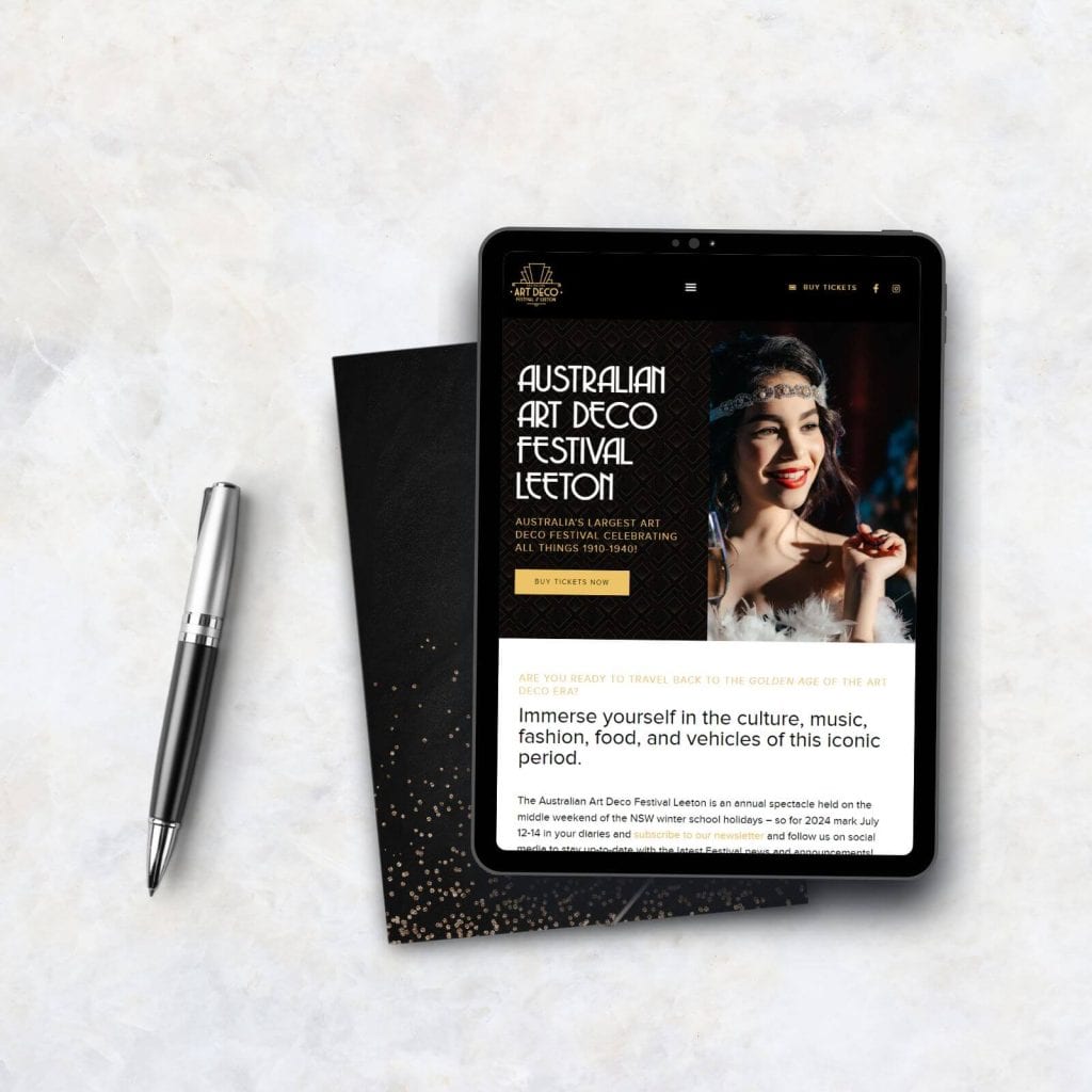 A tablet displaying the Australian Art Deco Festival Leeton website sits on a marble surface next to a closed black notebook and a silver pen.