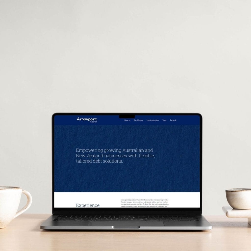 A laptop on a light wooden table displays a website for Arrowpoint, offering tailored debt solutions for Australian and New Zealand businesses. Two mugs are placed beside the laptop.