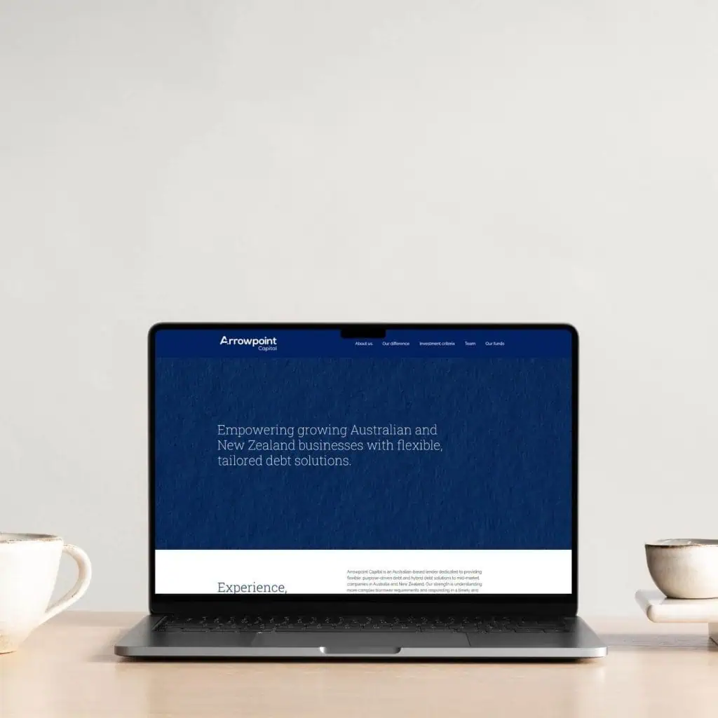 A laptop on a light wooden table displays a website for Arrowpoint, offering tailored debt solutions for Australian and New Zealand businesses. Two mugs are placed beside the laptop.