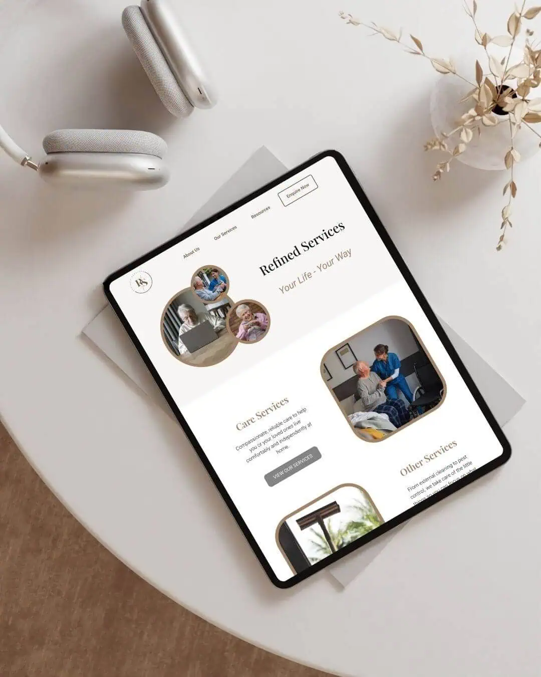 A tablet displaying a website for care services rests on a white table next to wireless headphones and a decorative vase with dried flowers.