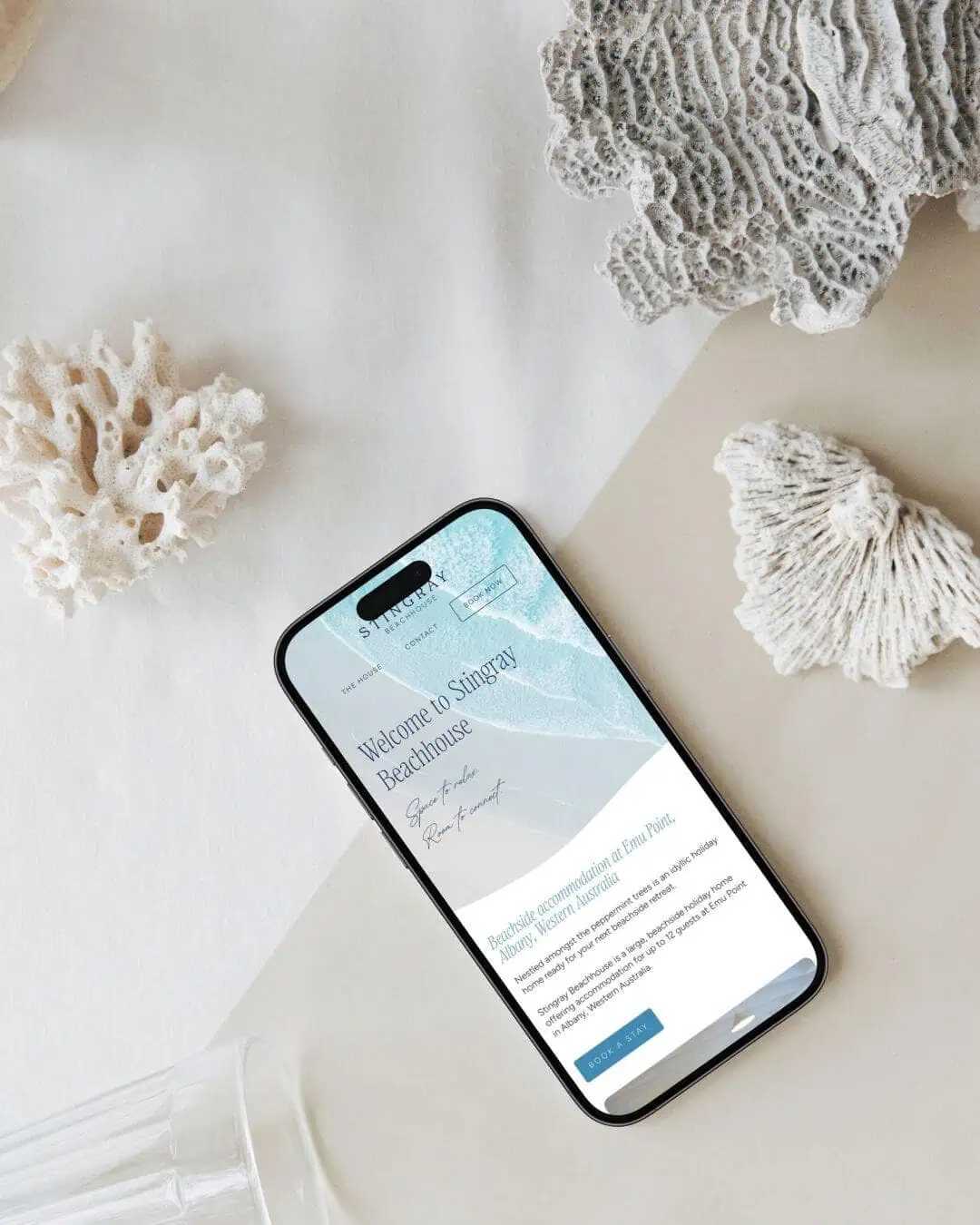 A smartphone displaying the Stingray Beachhouse website lies on a white surface next to pieces of coral and an empty glass.