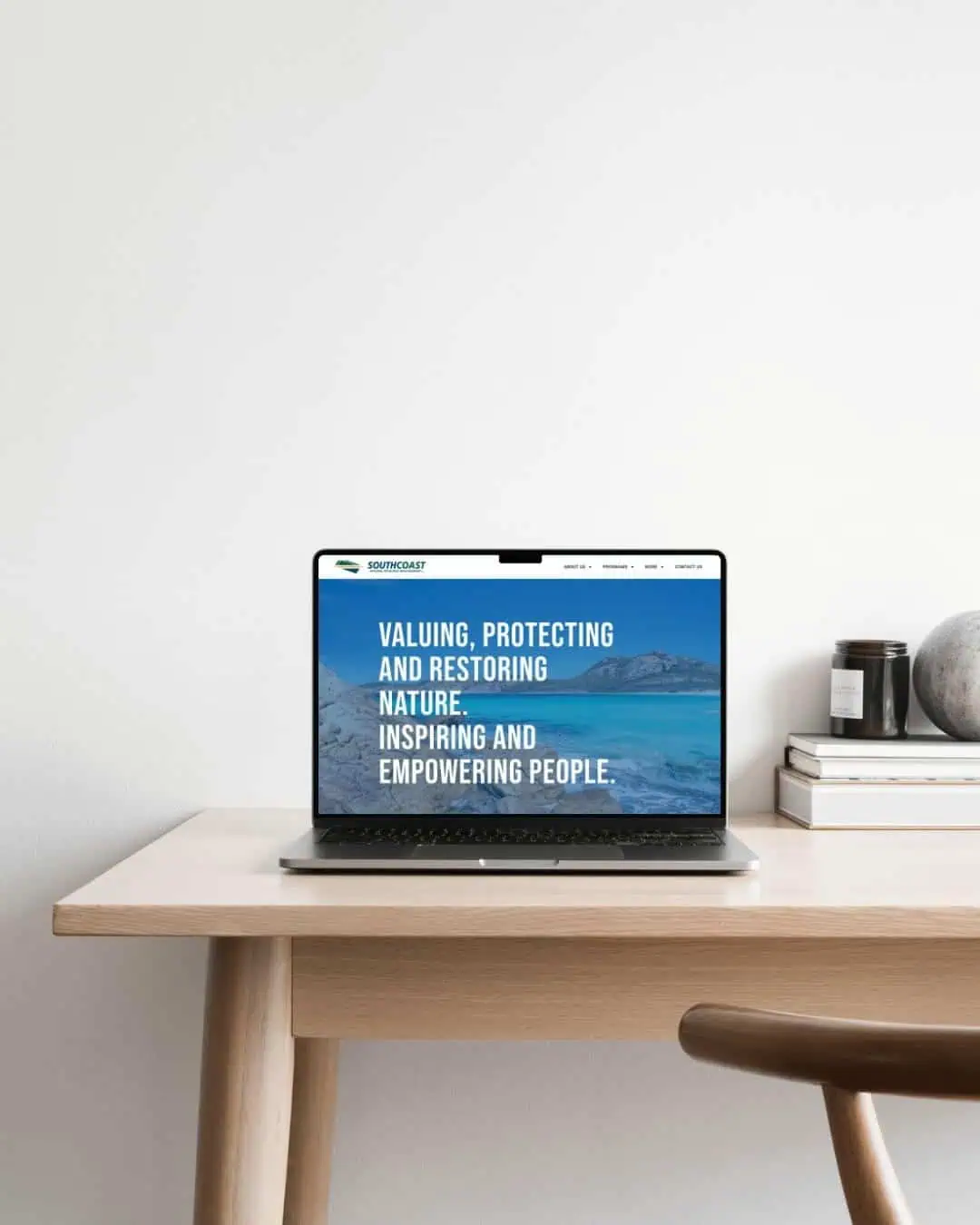 A laptop displaying a website with a nature-themed banner sits on a light wooden desk, next to a notebook, a pen, and two decorative objects against a plain white wall.