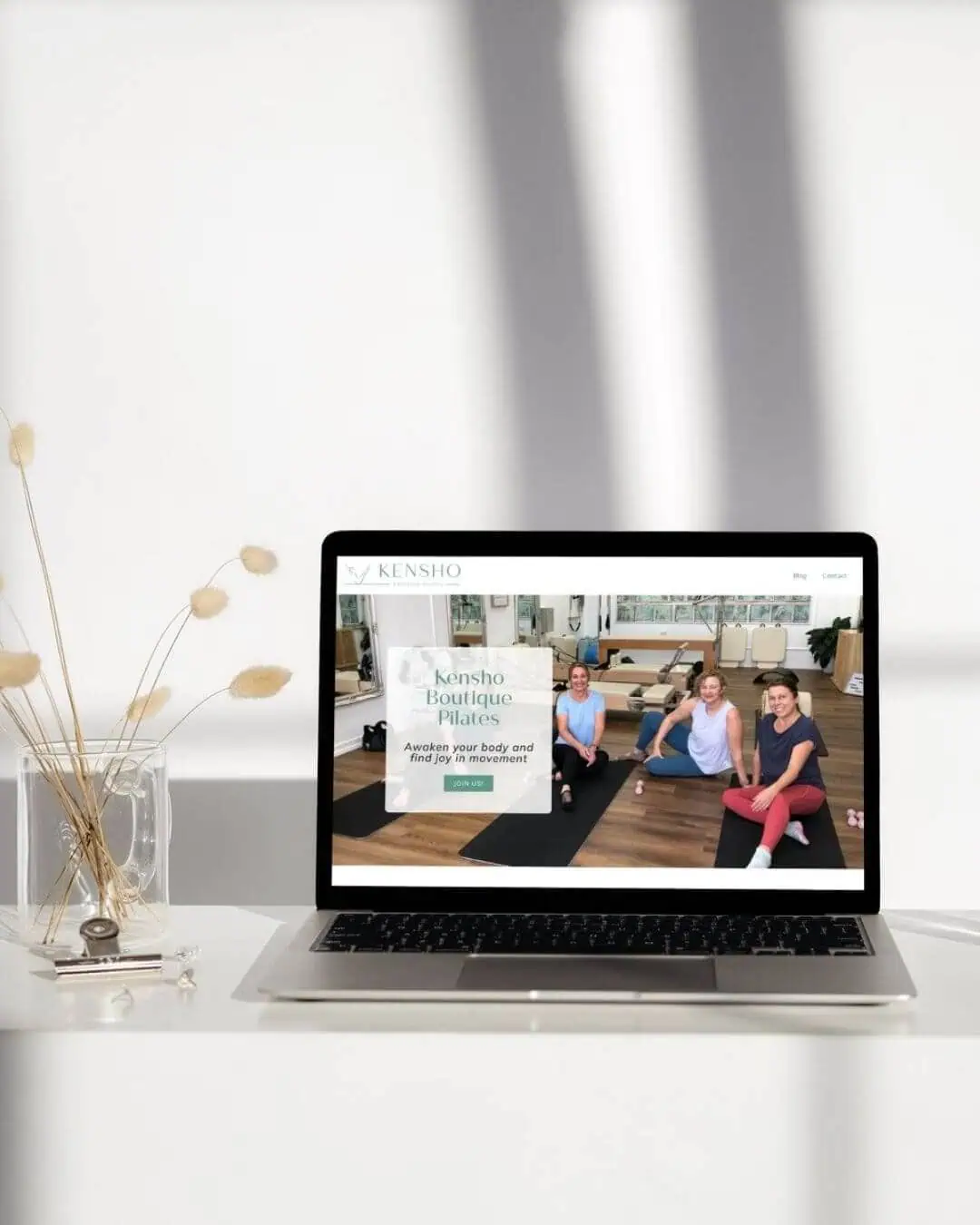 A laptop on a white table displays a website with three women doing Pilates on yoga mats. A glass vase with dried plants is beside the laptop. Light and shadows fall across the scene.