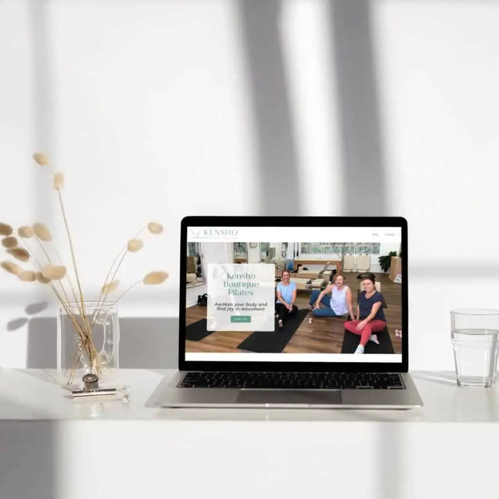 A laptop on a white desk displays a Pilates studio website. The desk also holds a glass of water and a vase with dried flowers. Sunlight casts window shadows on the wall.