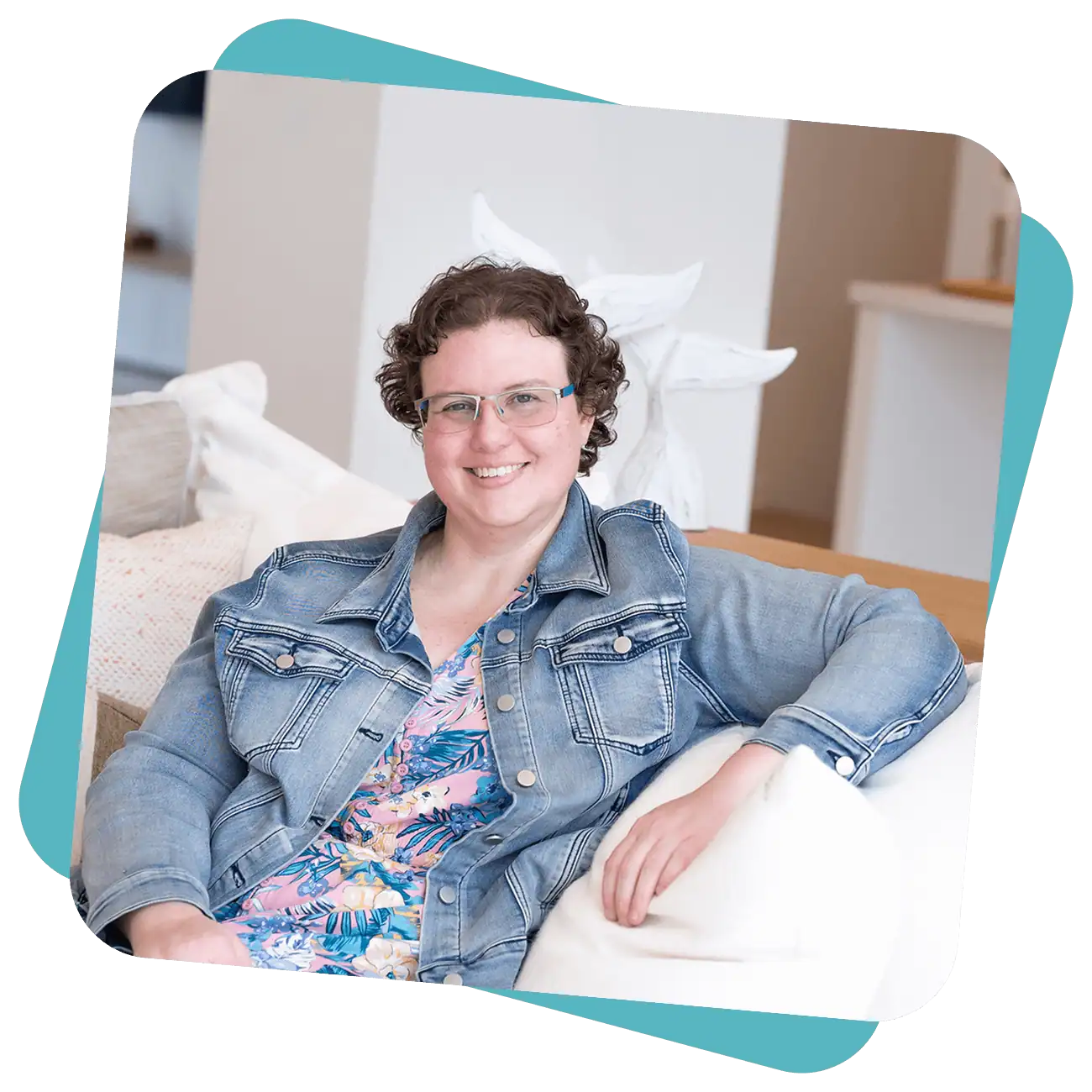 A person with short curly hair and glasses sits on a white sofa, wearing a denim jacket over a floral top, and smiling at the camera.