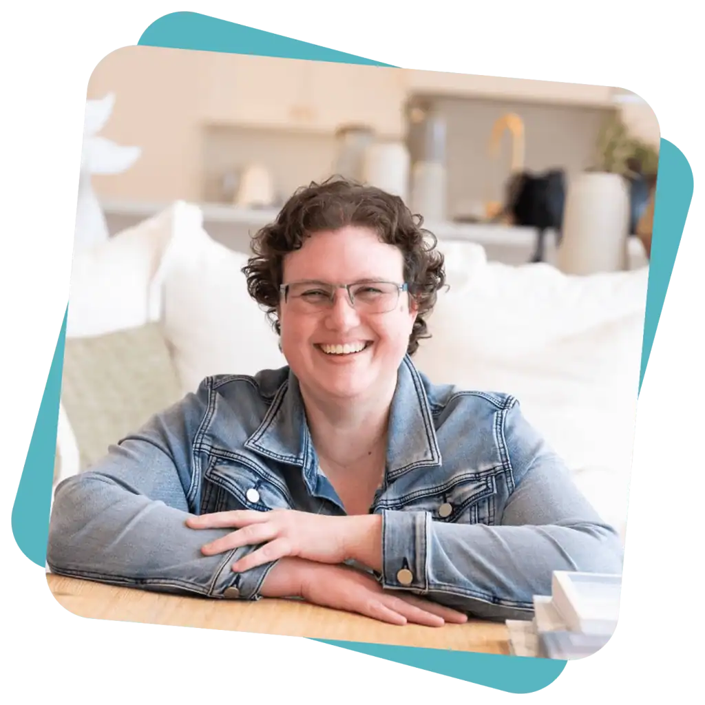 A person with short curly hair and glasses, wearing a denim jacket, smiles whilst sitting at a table in a bright, modern room.