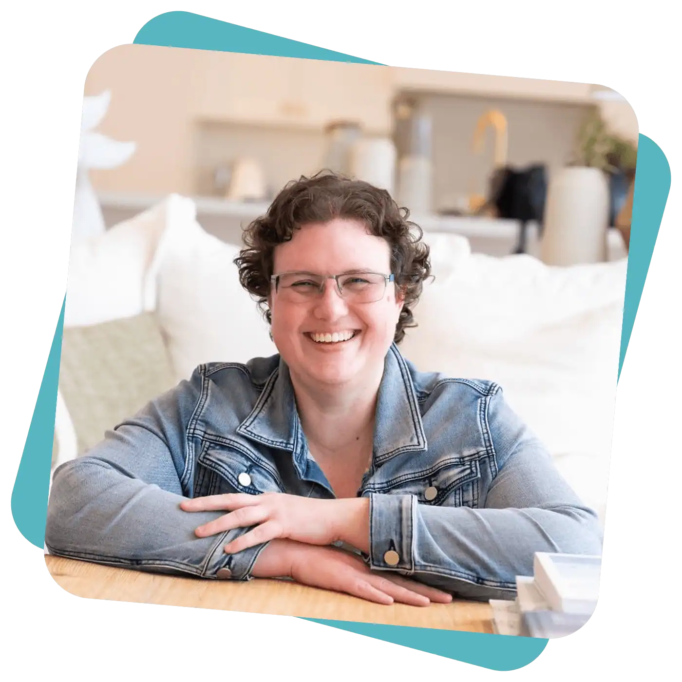 A person with short curly hair and glasses, wearing a denim jacket, smiles whilst sitting at a table in a bright, modern room.