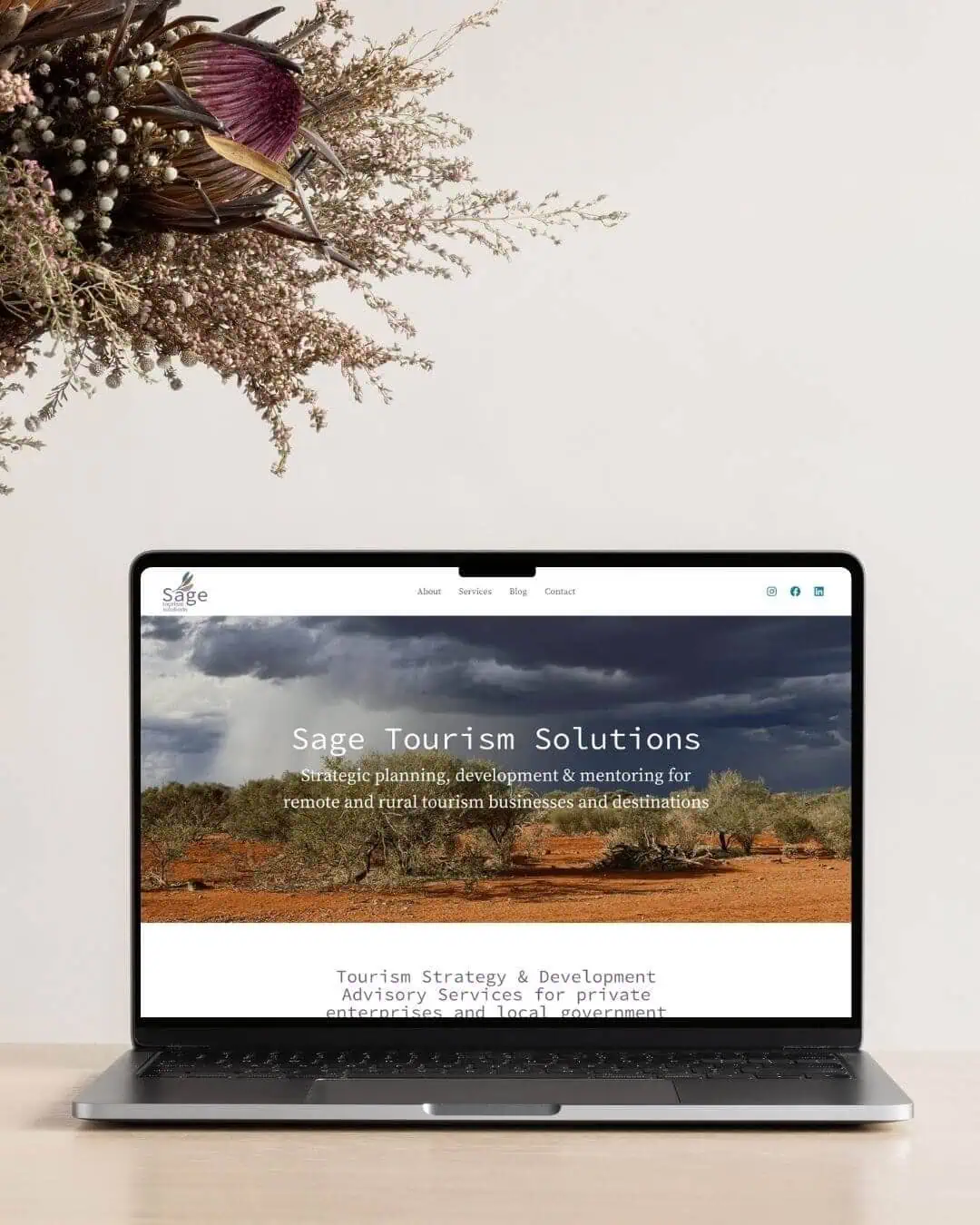 A laptop on a beige desk displays a website for Sage Tourism Solutions, with dried flowers in a vase placed to the left of the laptop.