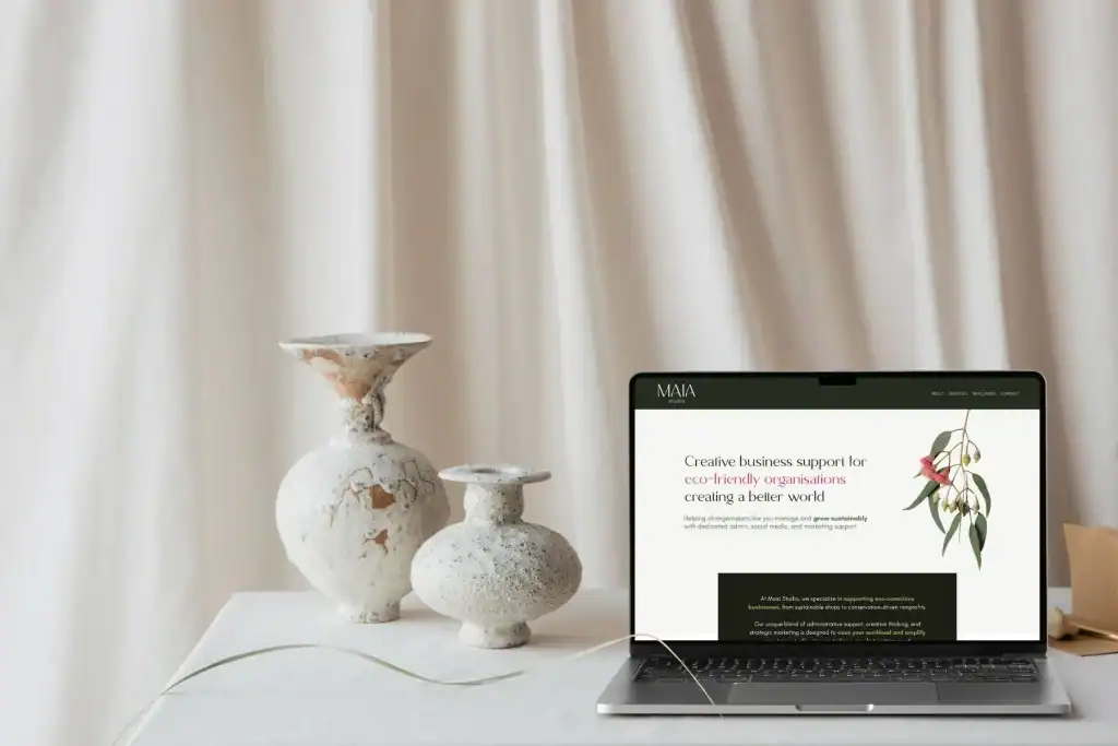 A laptop displaying a creative business support website sits on a white table next to three ceramic vases with a neutral curtain backdrop.