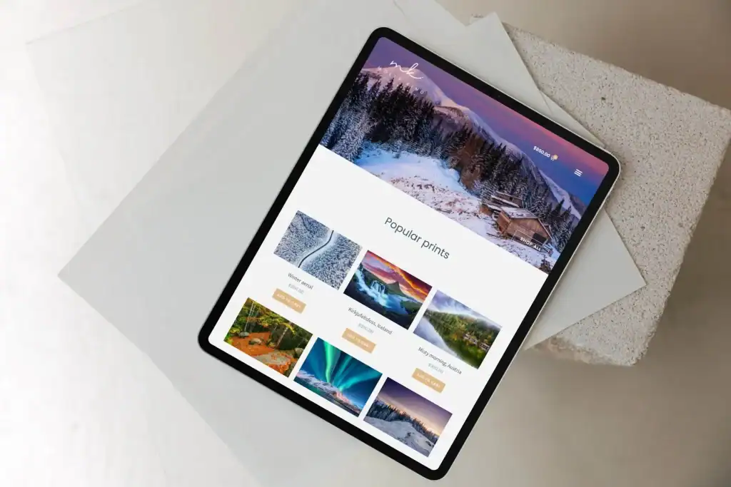 A tablet displaying a website with landscape photography prints, including categories for mountains, forests, auroras, deserts, and more. The tablet rests on white surfaces.