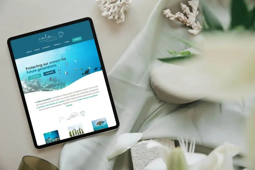 A tablet displaying a website about ocean conservation rests on a soft green fabric, surrounded by white flowers and coral pieces.