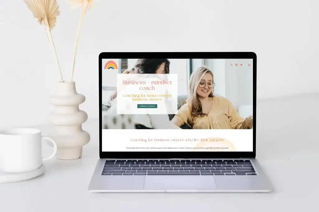A laptop on a white desk displays a website for a business mindset coach. A dried floral arrangement in a white vase is positioned to the left of the laptop.