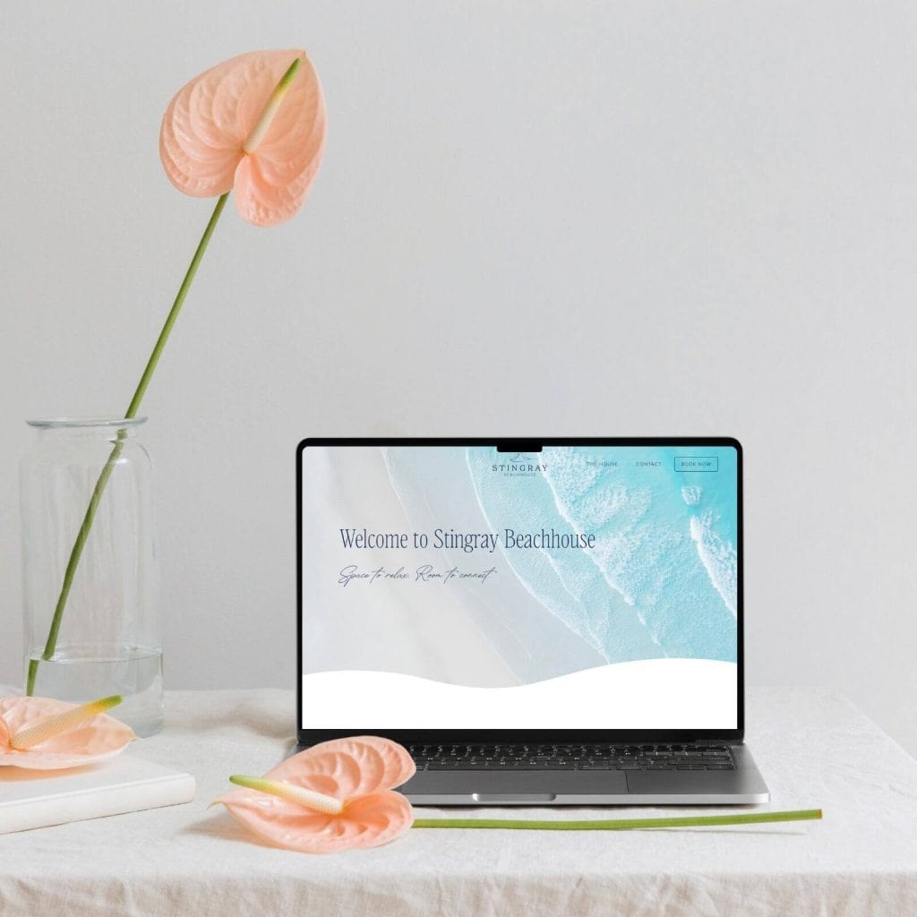 A laptop displaying a beachhouse website sits on a white table with pink anthurium flowers in a vase and on the table against a plain white background.