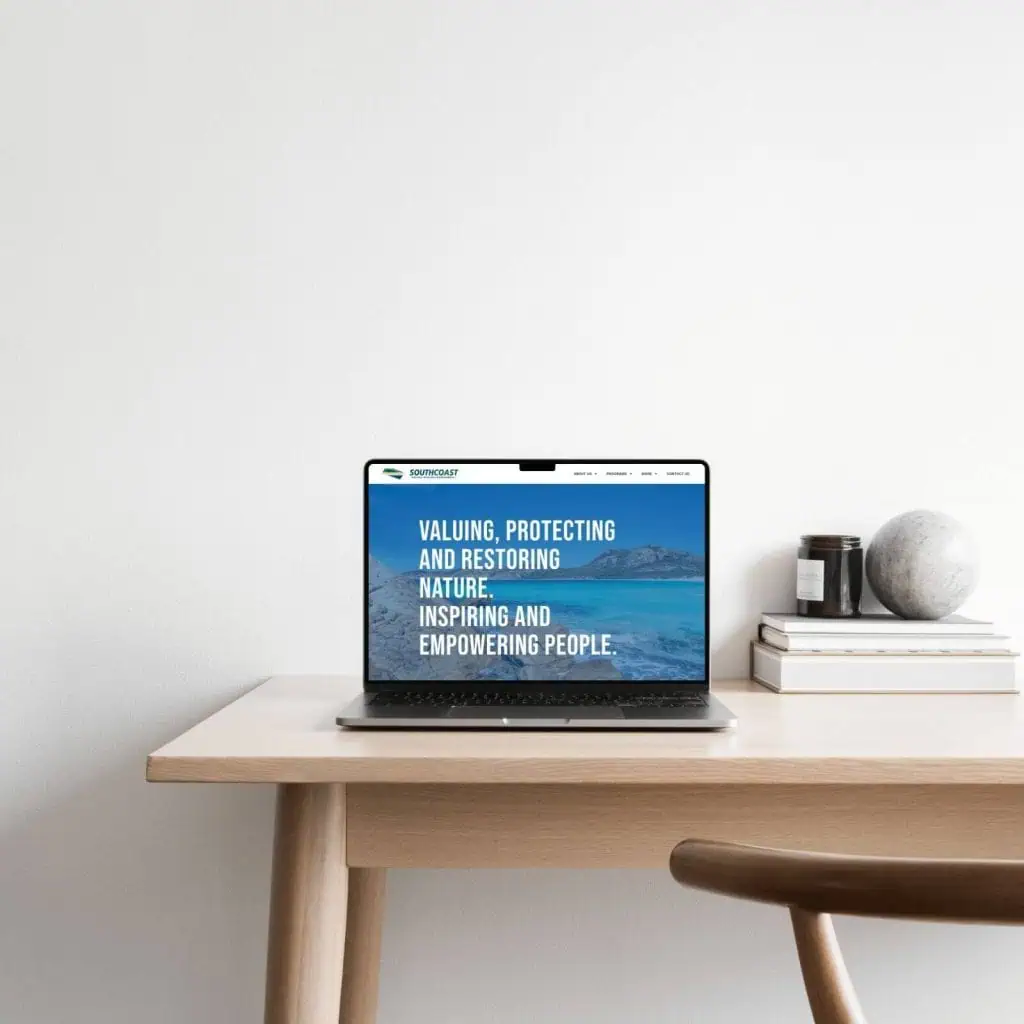 A laptop displaying a nature conservation website sits on a wooden desk next to stacked books, a marble sphere, and small containers, against a plain white wall.