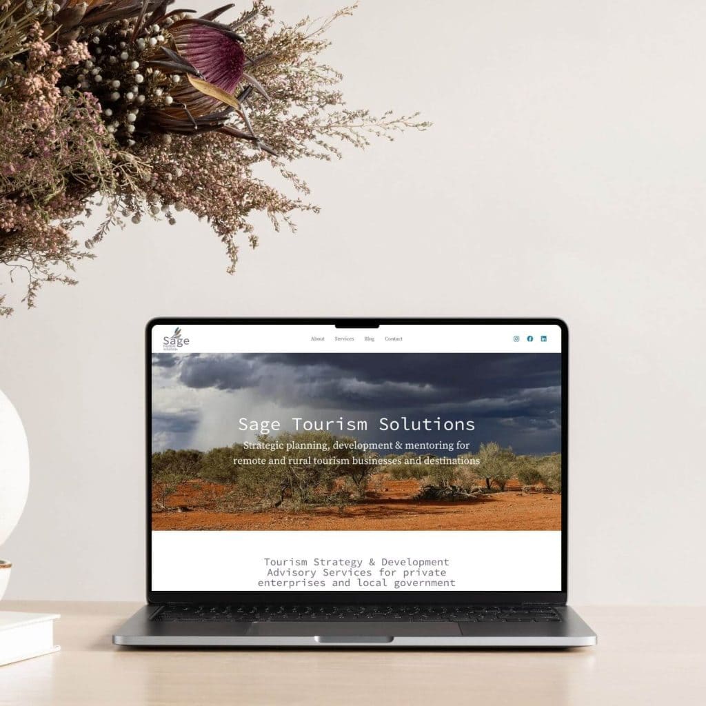 A laptop on a desk displays the Sage Tourism Solutions website, with a rural landscape image on the screen; a vase of dried flowers is positioned beside it.