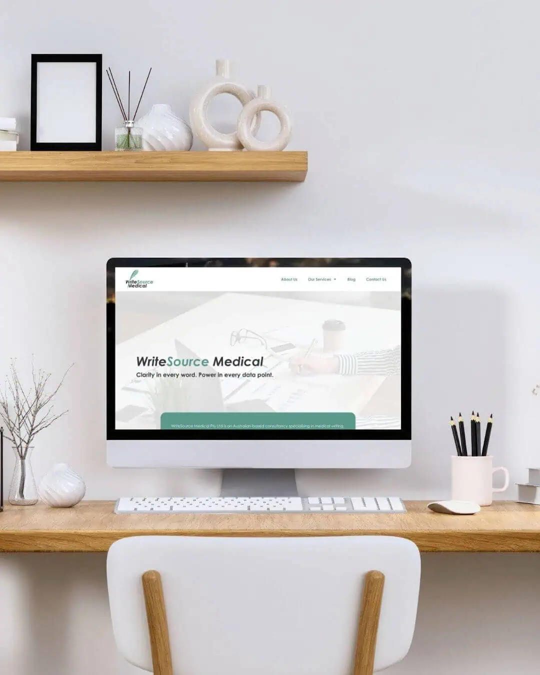 A computer on a wooden desk displays a website called WriteSource Medical. The workspace includes shelves with decor items, a mug, and a notepad.